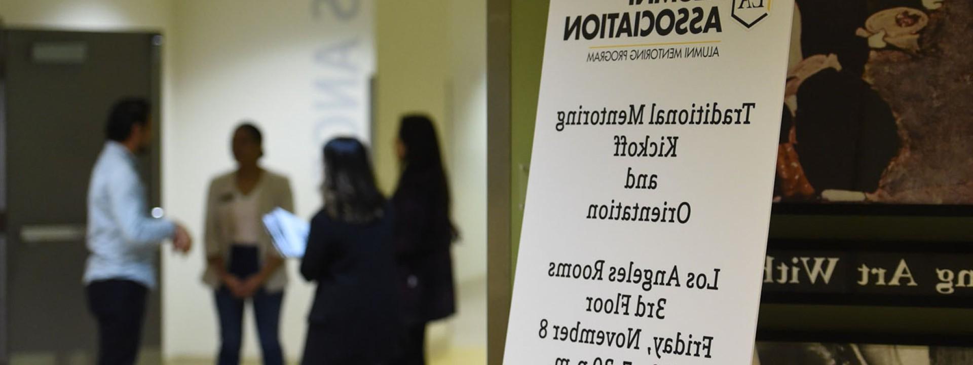 Sign for mentoring orientation with a group of people chatting in the background